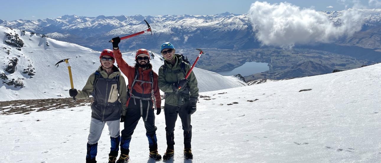 three students in snow gear holding mountaineering tools standing on snow with mountains and sunny clouds in the background