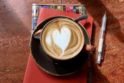 Artful cappaccino with heart-shaped foam, red notebook, pencil, sunny café table. 