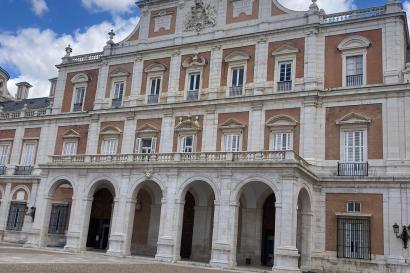 The face of the royal palace of Aranjuez, made of red brick and white arches. One Spanish flag flies above the left side of the facade.