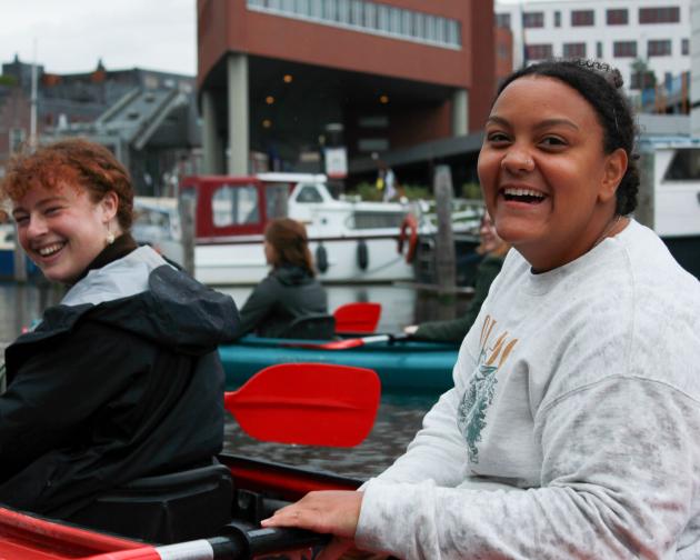 two students sitting in a kayak on a canal smiling and laughing with ores in hands in front of boats and buildings