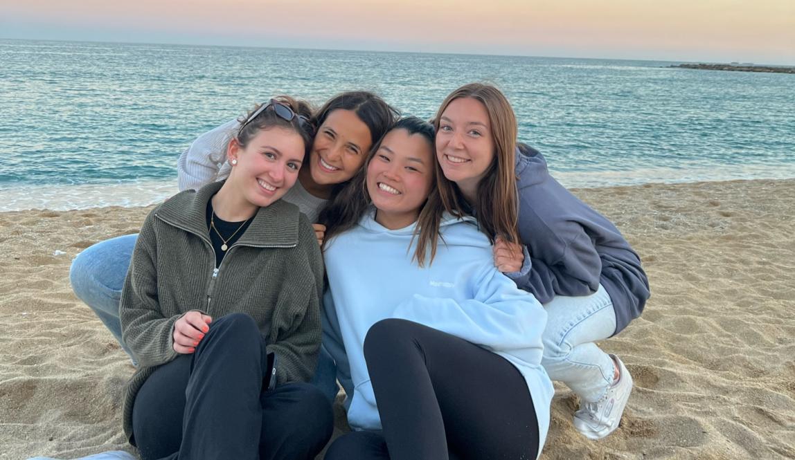 four students sitting on the sand at the beach with the ocean and the sunset in the background
