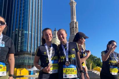 Two girls smile in front of a fountain and a statue. They are wearing bibs for a running race and medals.