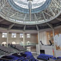 Inside of parliament building, with aluminum Eagle on window in the bottom right and blue chairs in a half circle, massive window dome with spike on ceiling