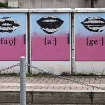 Three electrical boxes that have been spray painted, fading from light blue to purple, on each box there are lips in different positions mouthing out the word VAG 