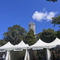 There are four white tents shown in the foreground. The tents are advertising university clubs such as tennis club and tramping club. In the background, there are trees and a white clock tower. The sky is bright blue with one white puffy cloud. 