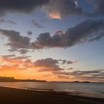 In this photo, the sun is setting, creating a bright orange sky with dark clouds. There is also a beach shown with calm waters.