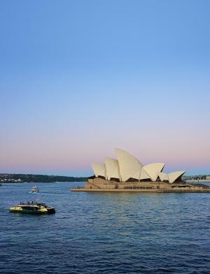 An aerial view of a boat sailing across Sydney Harbour in front of the Opera House at dusk