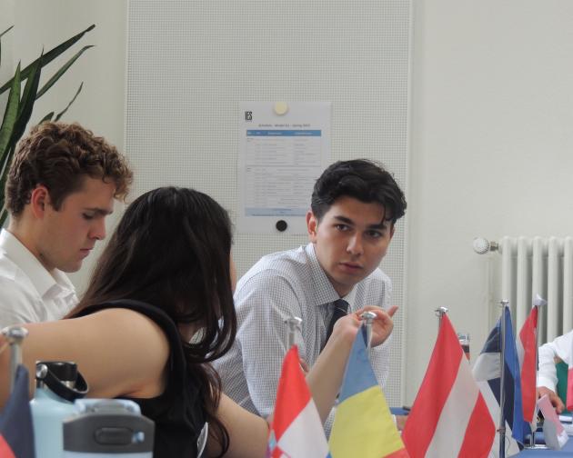 Three students look focused as they sit in class. On the table in front of them are different nation's flags.