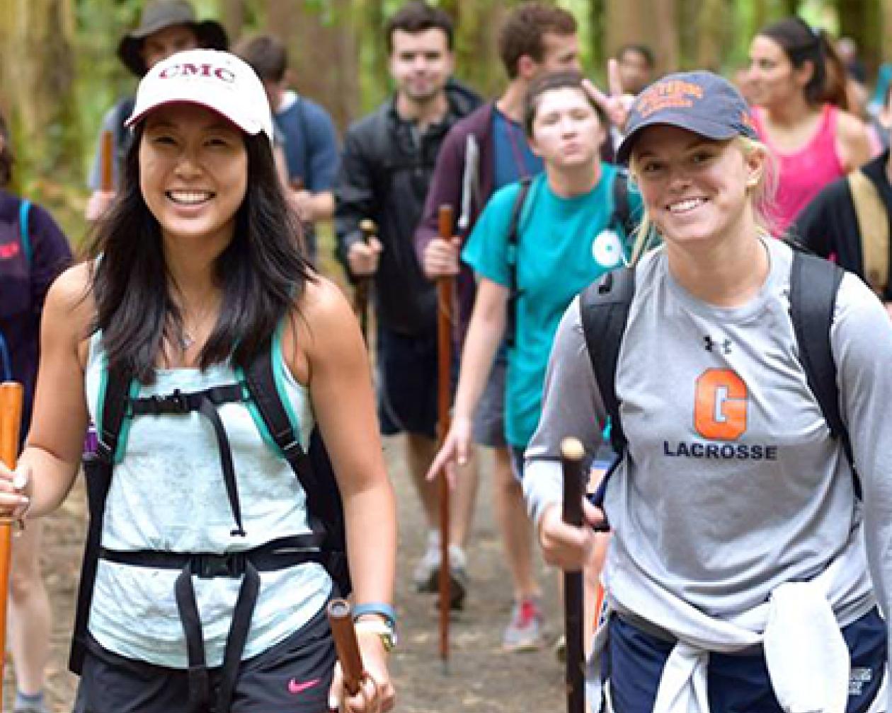 Students walking along a forest trail with walking sticks and smiling at the camera.