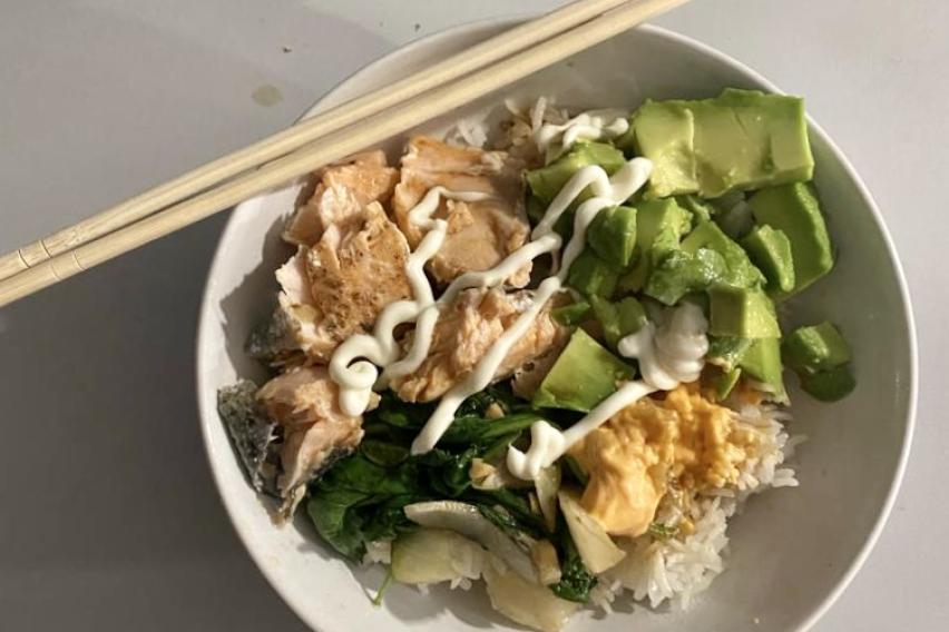 A bowl with chopsticks leaning on it, filled with fish, avocado, rice, and sauces. 