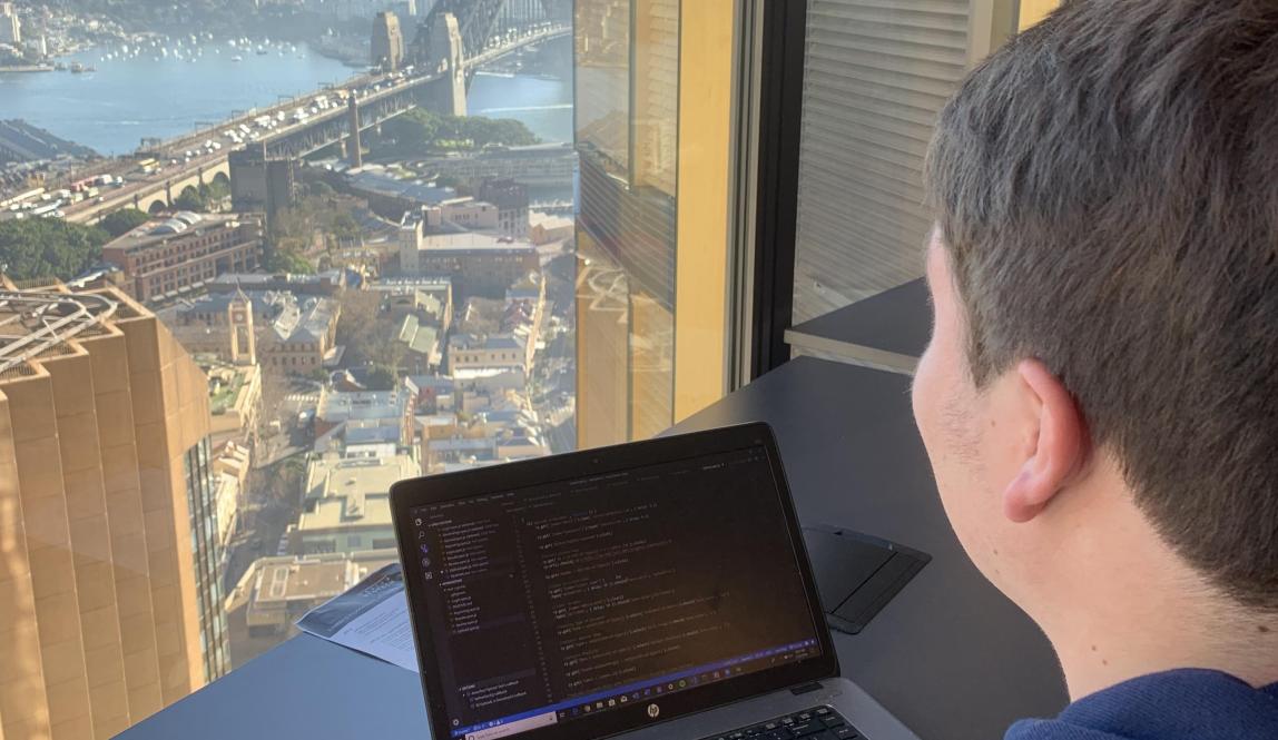 a student intern works on their computer while looking out the window at the Sydney Bridge