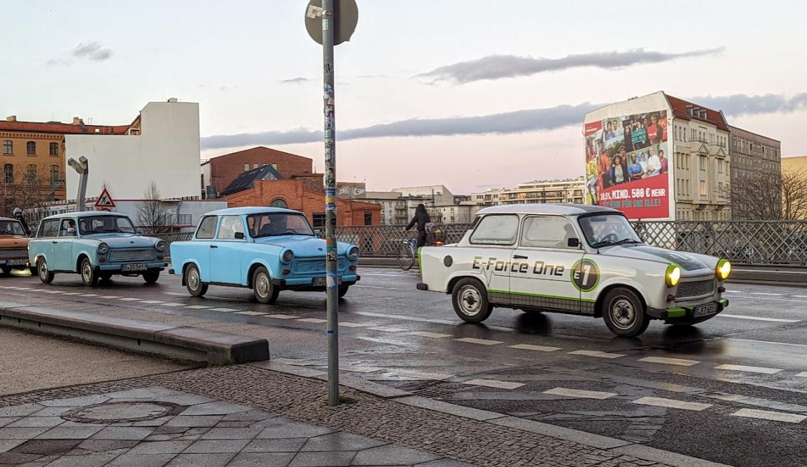 Three Trabi cars in street, two blue on the left and one white one on the right that says E-Force 1