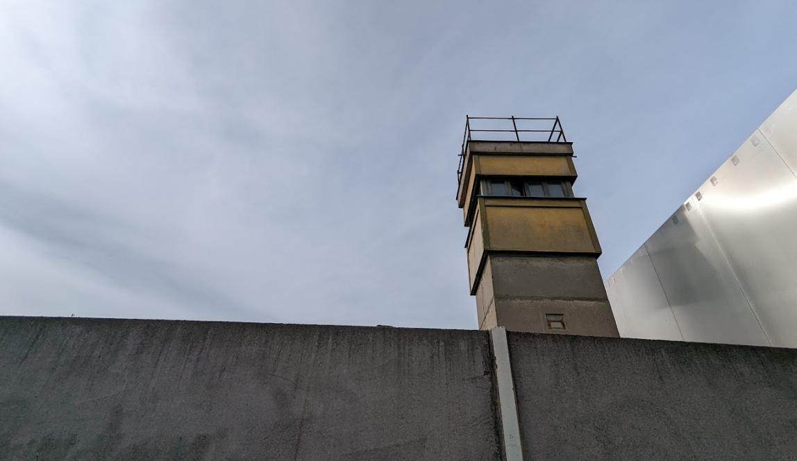 Concrete wall on bottom of picture with concrete guard tower behind it on the right, a metal wall sits behind the tower