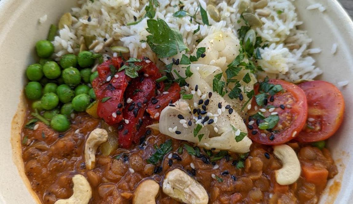 Paper bowl with white rice on one half, and orange/rust-colored lentil curry on the other half, the curry has green peas, cashews, tomatoes, and ginger slices on top of it