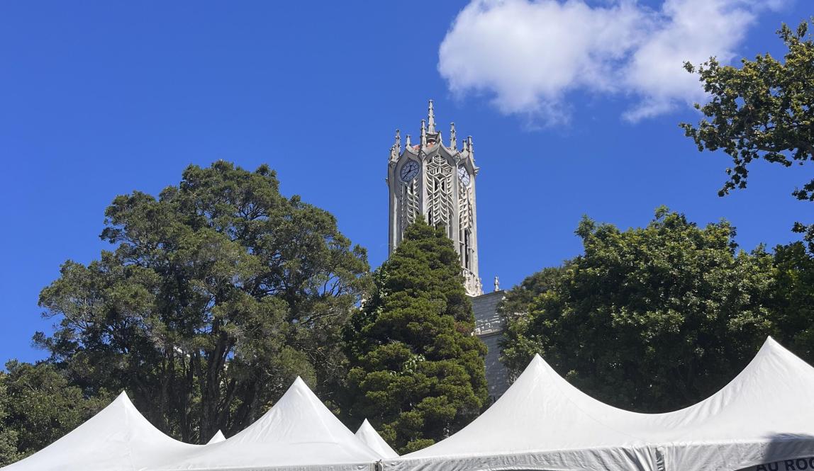 There are four white tents shown in the foreground. The tents are advertising university clubs such as tennis club and tramping club. In the background, there are trees and a white clock tower. The sky is bright blue with one white puffy cloud. 