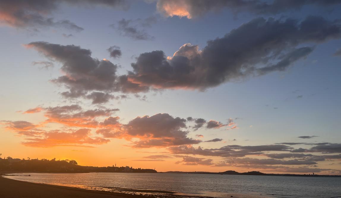 In this photo, the sun is setting, creating a bright orange sky with dark clouds. There is also a beach shown with calm waters.