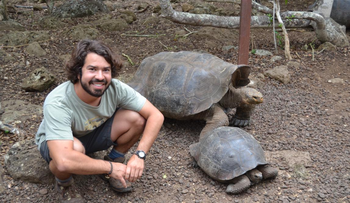 a student poses for a photo crouched down next to a giant tortoise and another smaller tortoise