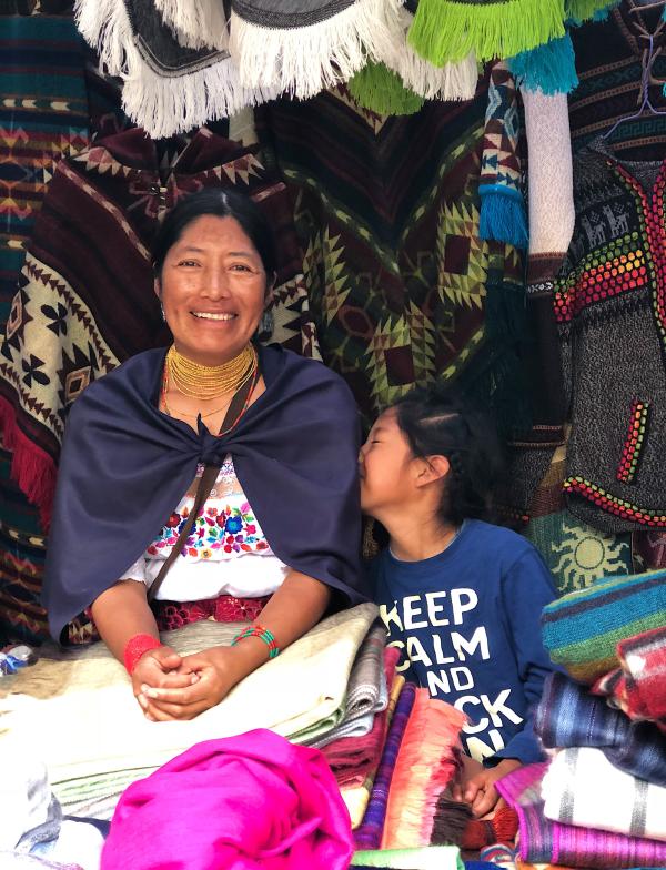 A person wearing traditional Otavalo wear (white shirt, shawl, and skirt) sits with a child wearing a "Keep Calm and Rock On" t-shirt. They are surrounded by handmade quilts and are smiling.