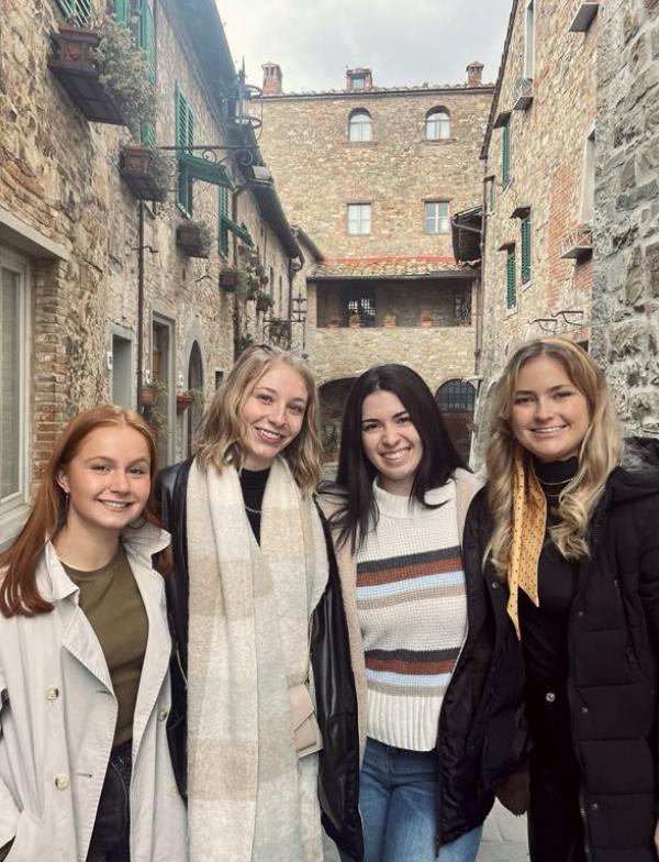 Students standing and smiling at the camera in a narrow street in Chianti, Italy. The buildings behind and around them are made of varying colors of brick.