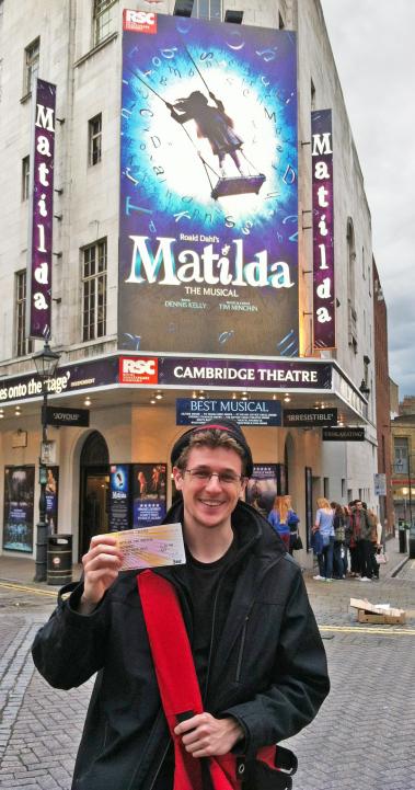 a male student standing outside of Cambridge Theatre with tickets to see the Matilda musical
