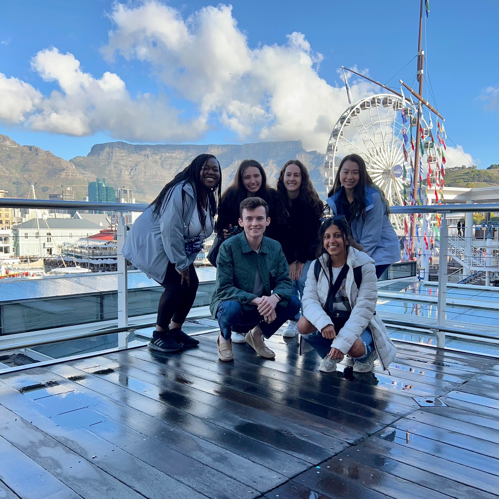 students posing for a picture on a pier. in the background, there is a Ferris wheel and mountains.