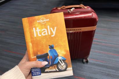Image of a hand holding up an orange guidebook titled Italy with a maroon suitcase in the background.