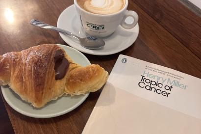 A cappuccino in a mug is sitting on a plate on top of a wooden table. Below it is a chocolate cornetto and next to it is Henry Miller's Tropic of Cancer book.