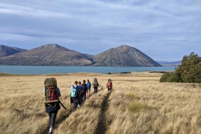 A group of people with backpacks walk down a trail across a plain, with a lake and mountains in the background.