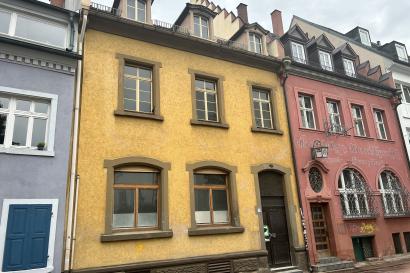 a snapshot of three buildings on a street in Freiburg, Germany. The one in the left corner is blue, the center one is yellow, and the one on the right is dusty red. A sliver of sky borders the top of the photo.
