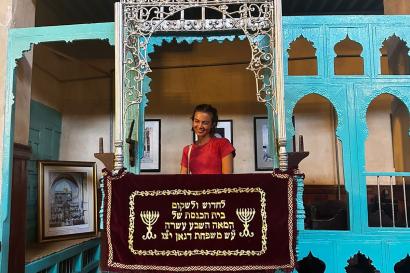 Mariama stands in the Bima of Ibn Danan Synagogue in Fes, Morocco. She is wearing a pink dress and smiling widely.
