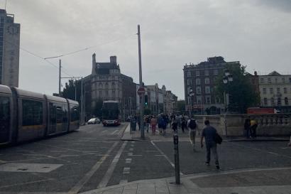 a view of Dublin, Ireland, from the street facing south. A tram train runs to the left, a building in the middle, and another building to the right. People walk towards the center from the right side. It is daytime and cloudy.