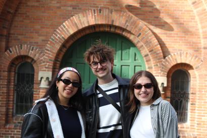Three students smile and stand in front of a green wooden door in a red brick building