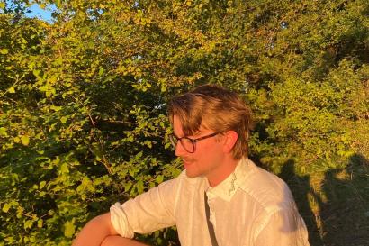 Boy sits on green bank in sunset lighting. He wears a white shirt and his pants are multicolored