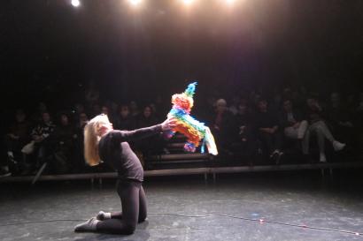 A girl kneels onstage, lifting a brightly colored piñata into the air while the audience looks on. 