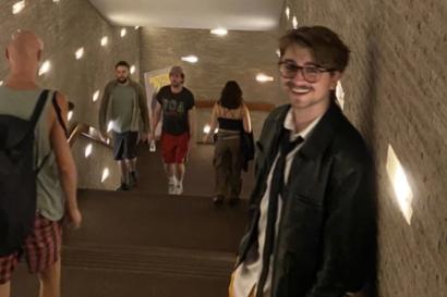 Boy wearing leather jacket, orange cargo shorts, black glasses, white shirt, and black tie stands in staircase with lights around it