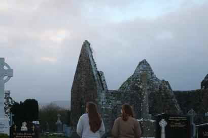Two people stand with their backs to the camera in lowlight, with the ruins of a cathedral and cemetery in the background.