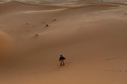 A view down over the desert, the dunes fade into the distance. Mari rides a snowboard down near the base of one dune.