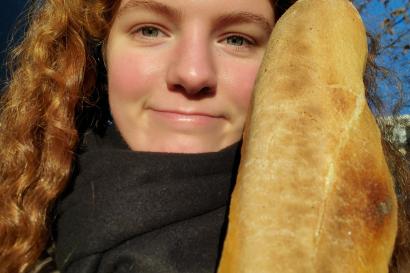 Ginger woman walking down the street smiling at the camera while holding a baguette in one hand.