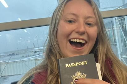 Maple Buescher holds her passport and smiles. Behind her, a jetbridge and airplane are visible through a window. Maple is white with blonde hair and wearing a teal jacket.
