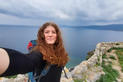 Ginger lady smiling into a camera at the top of a hill in front of the Monemvasia sea.