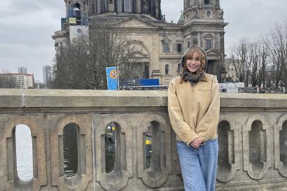 A girl poses in front of the Berlin Cathedral. She's wearing a grey scarf over her head, a tan jacket, jeans and black boots.