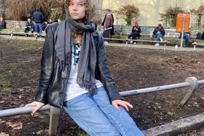 A girl sits on a railing in a park. She's wearing a gray scarf, black leather jacket, printed t-shirt, blue jeans and sneakers