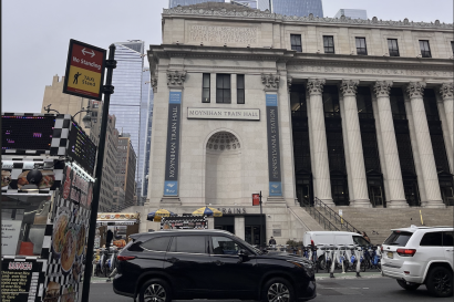 This photo shows the entrance of Moynihan Train Hall in New York City. The train hall is a large building supported by several columns and many stairs. It also has two decorative ribbons with engravings that say Moynihan Train Hall and Pennsylvania Station. 