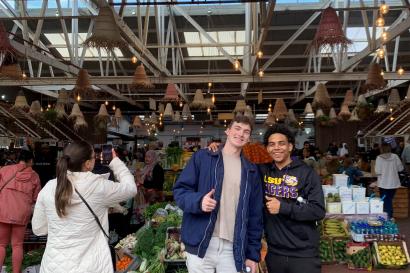 two students holding a thumbs up in front of a food market with signs 