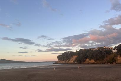 A large beach with three people and a cliff are shown. The sun is setting, creating a pale blue sky and pink clouds. 