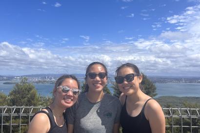 In this picture, there are three girls posing in front of the Auckland skyline. The sky has a few clouds, but is mostly clear. The girl on the left wears a blank tank top and reflective sunglasses. The girl in the middle wears a grey t-shirt with black sunglasses. The girl on the right also wears a black tank top with black sunglasses.