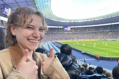 A young girl gives a thumbs-up in front of a Hertha BSC soccer game