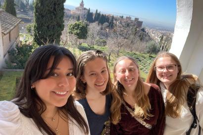 Four girls smile at the camera in a selfie. Behind them a valley with an old castle is visible in front of a blue sky.