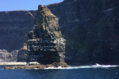 A tall rock formation rises from the ocean near towering cliffs under a blue sky. Peaceful waves crash against the stack's base.