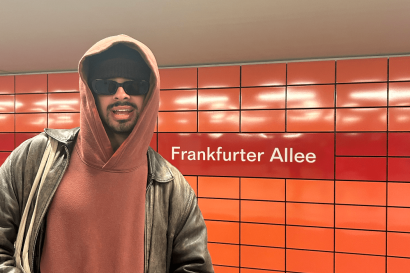 A photo of me in the underground with a bright orange and red backdrop including the words "Frankfurter Allee"